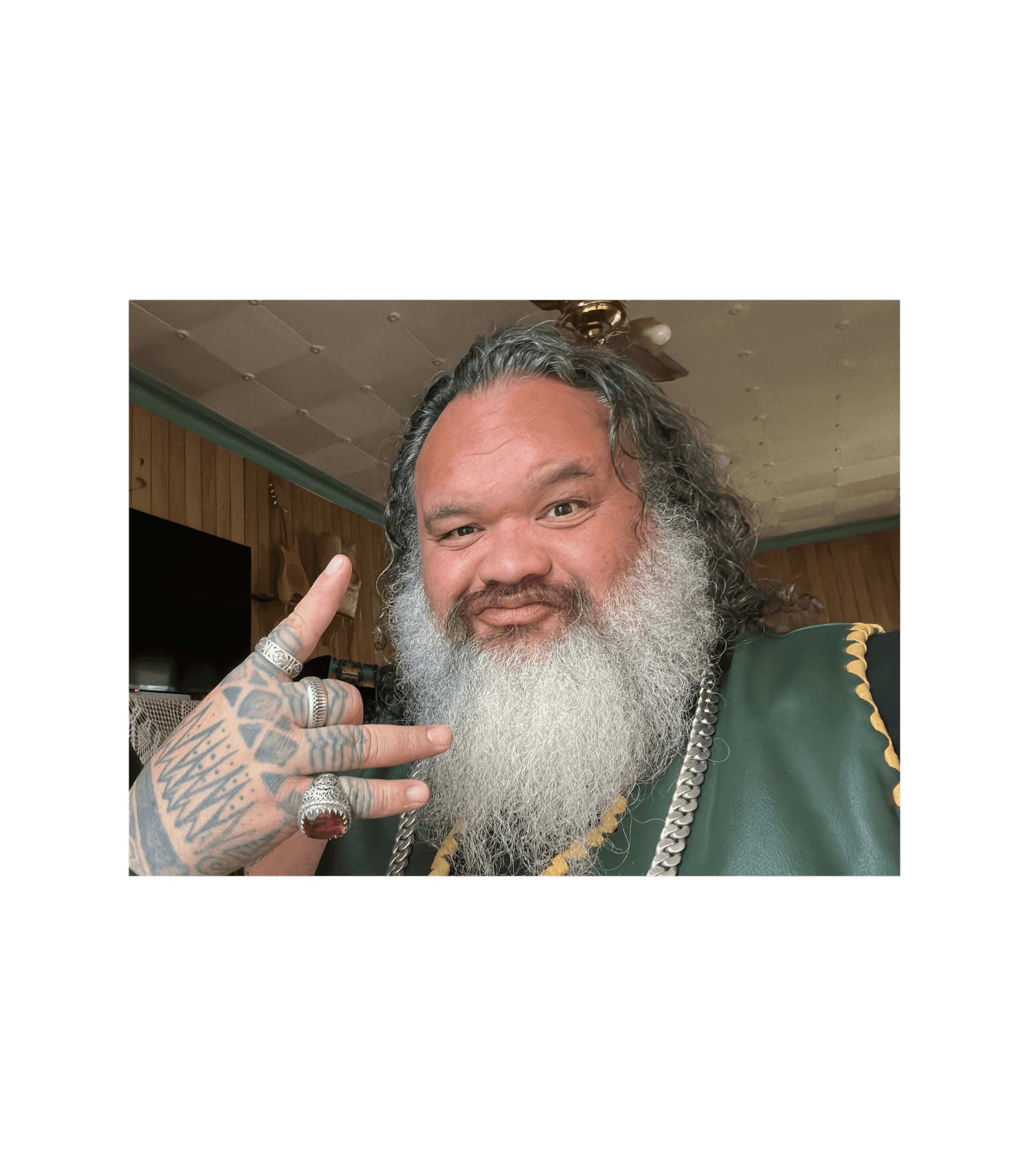 Man with a long beard and tattoos on his hand making a peace sign gesture while smiling indoors.